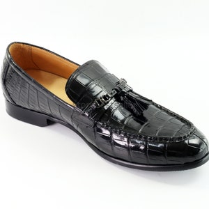 May include: Black leather loafers with a crocodile pattern and a tassel detail. The loafers have a metal buckle closure.