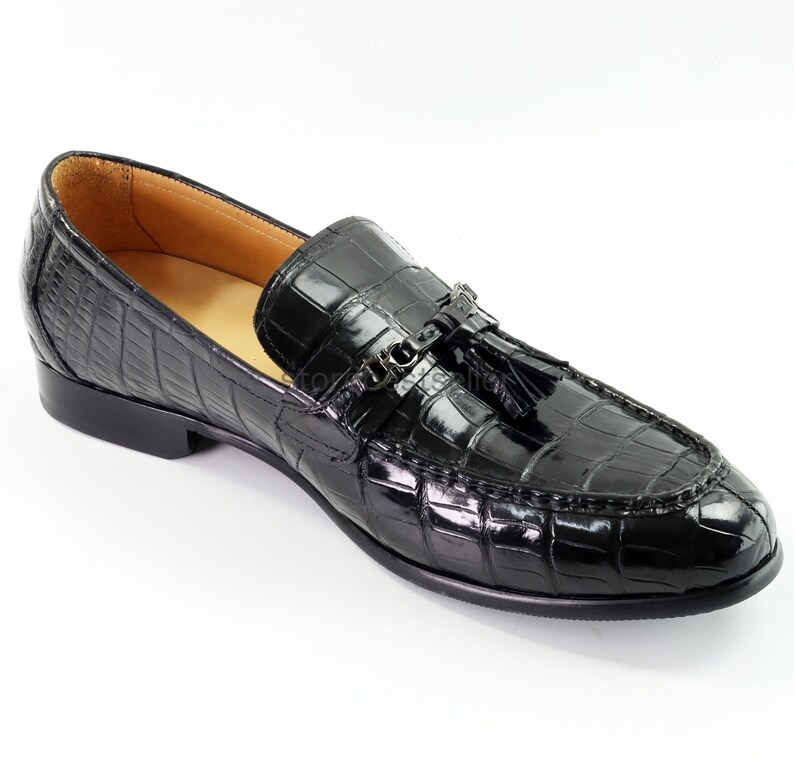May include: Black leather loafers with a crocodile pattern and a tassel detail. The loafers have a metal buckle closure.