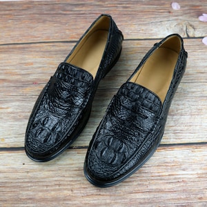 May include: A pair of black leather loafers with a crocodile skin texture. The shoes have a classic slip-on design with a rounded toe and a low heel. The interior is a light tan color.