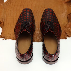 May include: A pair of red and black leather dress shoes with a crocodile pattern. The shoes have laces and are on a white background.
