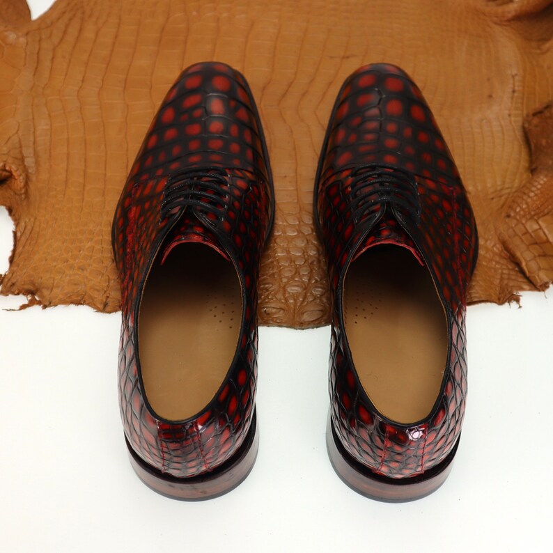 May include: A pair of red and black leather dress shoes with a crocodile pattern. The shoes have laces and are on a white background.