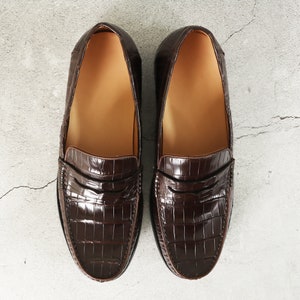 Puede incluir: Un par de mocasines de cuero marr&oacute;n con un patr&oacute;n de cocodrilo en relieve. Los mocasines tienen un dise&ntilde;o cl&aacute;sico de mocas&iacute;n penny con una sola correa en el empeine.