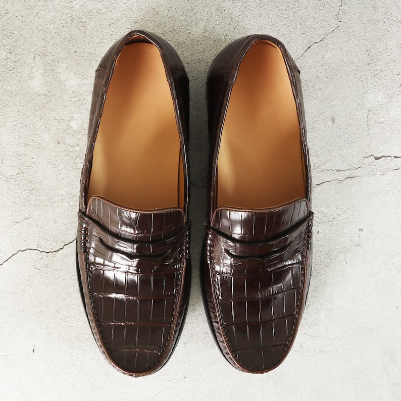 Puede incluir: Un par de mocasines de cuero marr&oacute;n con un patr&oacute;n de cocodrilo en relieve. Los mocasines tienen un dise&ntilde;o cl&aacute;sico de mocas&iacute;n penny con una sola correa en el empeine.