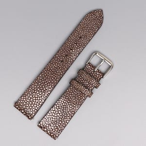 May include: Brown watch strap with a textured, speckled pattern. The strap is made of a material that resembles stingray skin. The buckle is silver-colored and rectangular. The strap is shown in two pieces, with one end featuring the buckle.