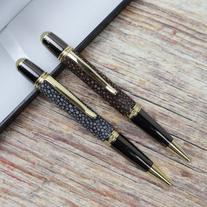 May include: Two black and gold pens with a textured, patterned design. The pens are lying on a wooden surface.