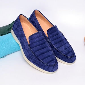 May include: A pair of navy blue loafers with a textured, crocodile-like pattern. The shoes have a rounded toe, a low profile, and a cream-colored sole. The interior is a light tan color. The shoes are displayed on a white surface.