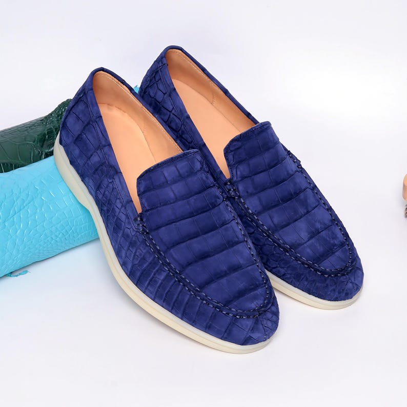 May include: A pair of navy blue loafers with a textured, crocodile-like pattern. The shoes have a rounded toe, a low profile, and a cream-colored sole. The interior is a light tan color. The shoes are displayed on a white surface.