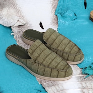 May include: A pair of olive green crocodile skin slip-on shoes. The shoes have a textured surface and a light beige sole. The background features blue and white leather textures.