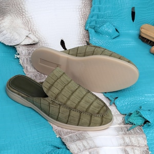 May include: Olive green leather slip-on shoes with a crocodile pattern. The shoes have a light beige sole and a dark green interior. The image shows the shoes on a blue and white textured surface.