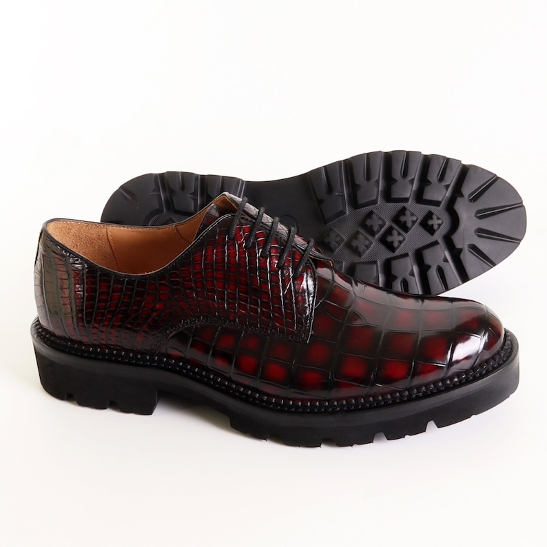 May include: A pair of burgundy leather lace-up shoes with a crocodile-embossed pattern. The shoes have a thick black rubber sole.