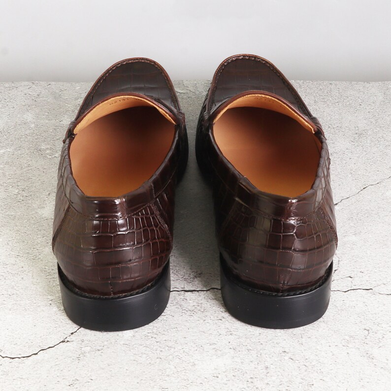 Puede incluir: Un par de mocasines de cuero marr&oacute;n con un patr&oacute;n de cocodrilo en relieve. Los mocasines tienen una suela de goma negra y un forro de cuero marr&oacute;n.