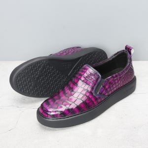 May include: A pair of slip-on sneakers with a textured, purple faux-crocodile skin pattern. The shoes have a black sole and a small pull tab at the heel. The second shoe is upside down, showing the black tread.