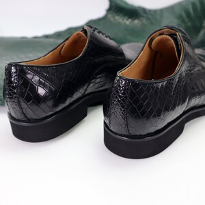 K&ouml;nnte beinhalten: Ein Paar schwarze Lederschuhe mit Kroko-Muster. Die Schuhe haben eine Schn&uuml;rung und eine dicke Sohle.