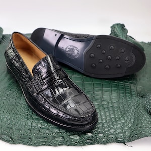 Puede incluir: Un par de mocasines de cuero negro con estampado de cocodrilo. Los mocasines tienen una suela de cuero con un patr&oacute;n texturizado. Los mocasines est&aacute;n sobre una superficie de piel de cocodrilo verde.