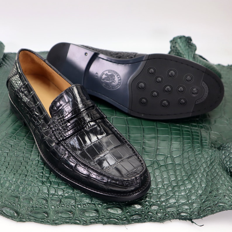 Puede incluir: Un par de mocasines de cuero negro con estampado de cocodrilo. Los mocasines tienen una suela de cuero con un patr&oacute;n texturizado. Los mocasines est&aacute;n sobre una superficie de piel de cocodrilo verde.