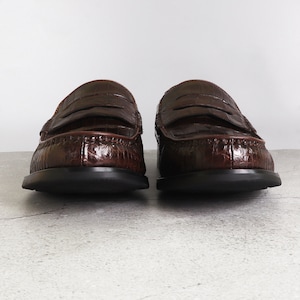 Puede incluir: Un par de mocasines de cuero marr&oacute;n con estampado de cocodrilo. Los mocasines tienen una suela de goma negra y una tira penny.