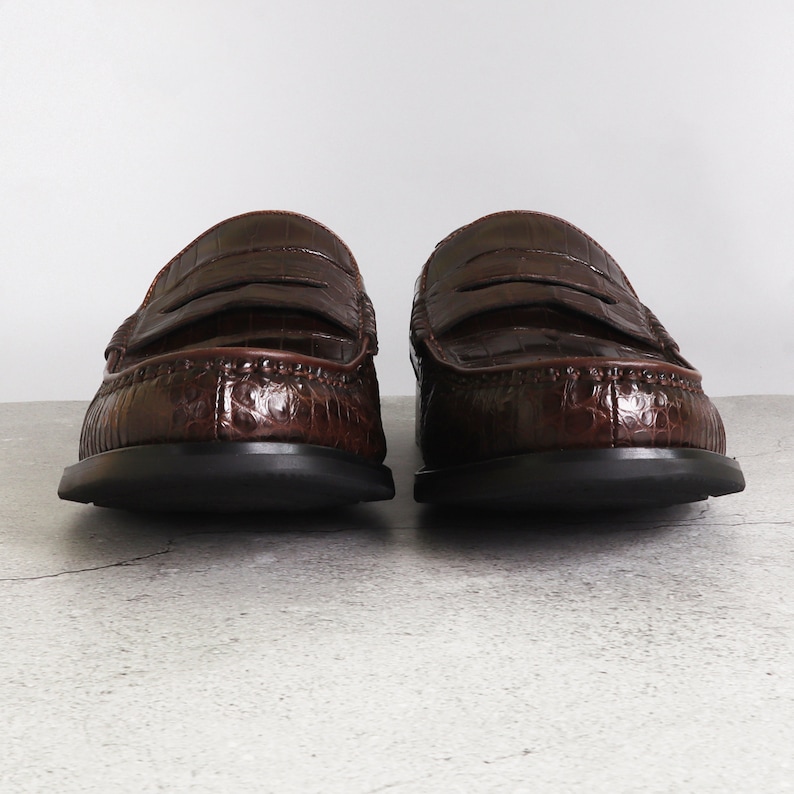 Puede incluir: Un par de mocasines de cuero marr&oacute;n con estampado de cocodrilo. Los mocasines tienen una suela de goma negra y una tira penny.