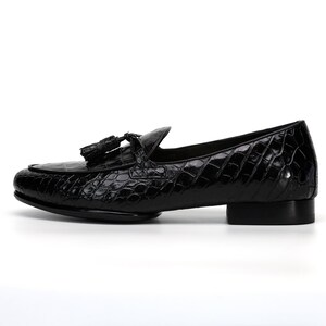 May include: Black leather loafer with a textured, crocodile-like pattern. The shoe features a rounded toe, a small heel, and a tassel detail. A classic, stylish footwear option.