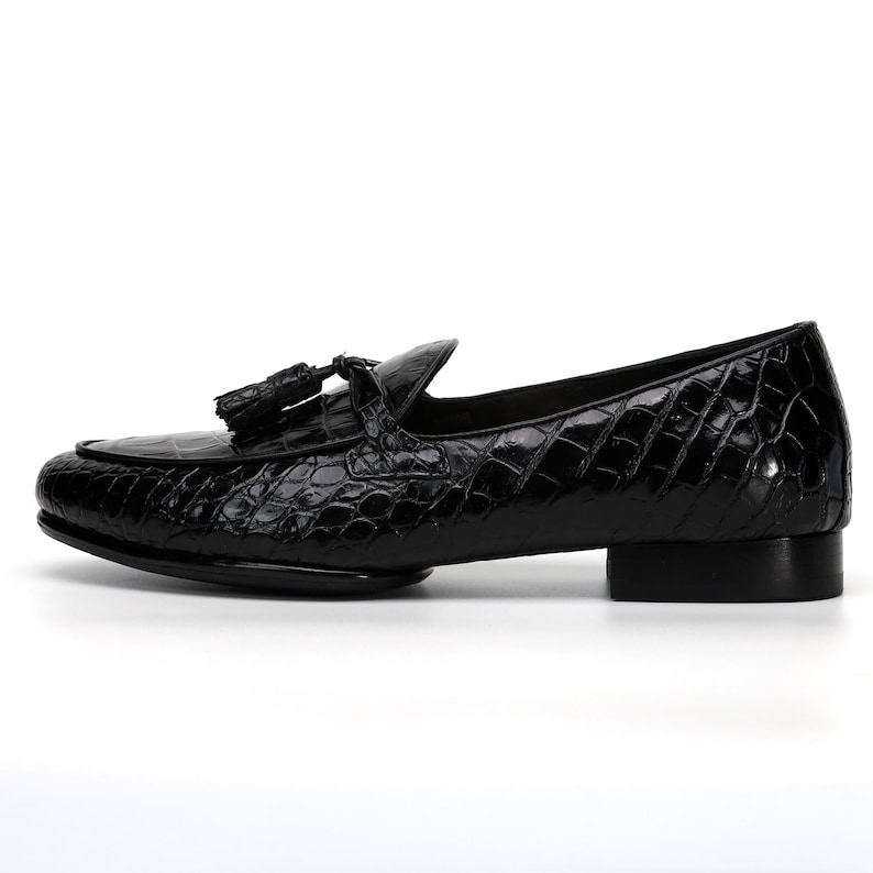 May include: Black leather loafer with a textured, crocodile-like pattern. The shoe features a rounded toe, a small heel, and a tassel detail. A classic, stylish footwear option.