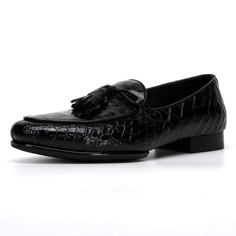 May include: Black leather loafer with a textured, crocodile-like pattern. The shoe features a tassel detail and a low heel. The design is sleek and classic, suitable for formal or semi-formal occasions.