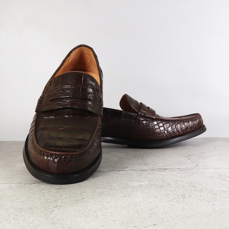 Puede incluir: Un par de mocasines de cuero marr&oacute;n con estampado de cocodrilo. Los mocasines tienen punta puntiaguda y un dise&ntilde;o de deslizamiento.