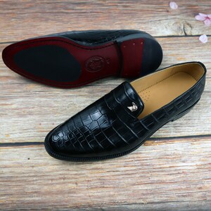 May include: A pair of black leather loafers with a crocodile pattern. The loafers have a silver buckle on the front and a brown leather sole.