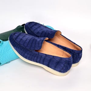 May include: A pair of blue suede loafers with a crocodile skin pattern. The shoes have a rounded toe and a cream-coloured sole. The loafers are displayed on a white surface.