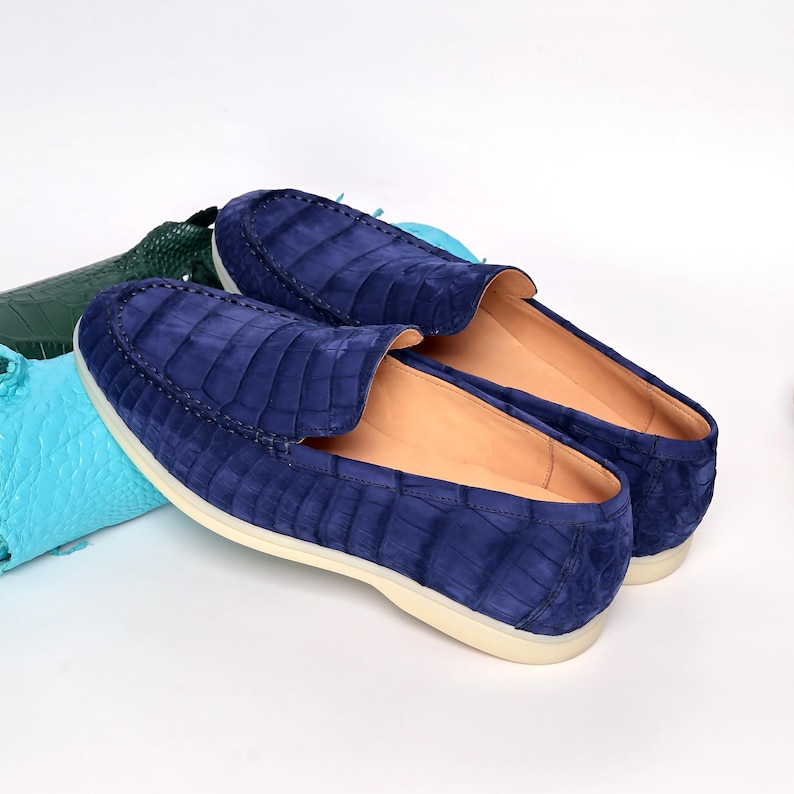 May include: A pair of blue suede loafers with a crocodile skin pattern. The shoes have a rounded toe and a cream-coloured sole. The loafers are displayed on a white surface.