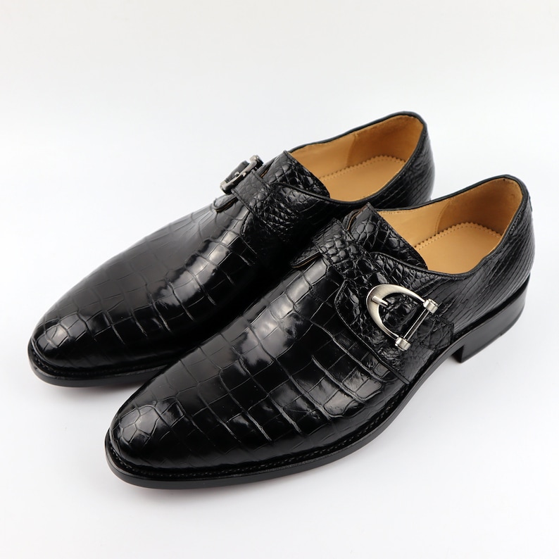 May include: A pair of black leather dress shoes with a crocodile pattern. The shoes have a single buckle strap with a silver buckle.