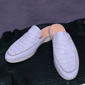 May include: A pair of light blue crocodile-textured leather slip-on shoes. The shoes have a rounded toe, a low heel, and a light-colored sole. The shoes are displayed on a dark green textured surface.