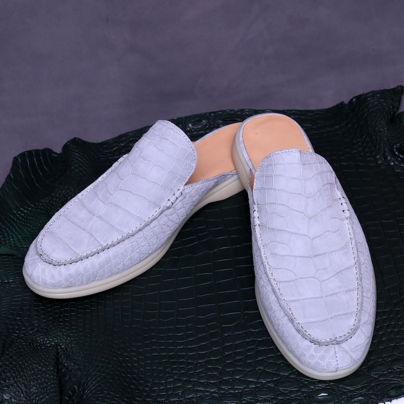 May include: A pair of light blue crocodile-textured leather slip-on shoes. The shoes have a rounded toe, a low heel, and a light-colored sole. The shoes are displayed on a dark green textured surface.