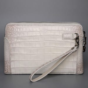 May include: A white clutch with a crocodile skin texture. The rectangular bag has a zipper closure and a wrist strap. The corners and edges of the bag have a darker, mottled pattern. A silver-colored clasp and zipper pull are visible.