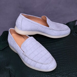 May include: A pair of light blue loafers with a crocodile skin texture. The shoes have a rounded toe and a cream-colored sole. The interior is a light tan color. The shoes are displayed on a dark green surface.