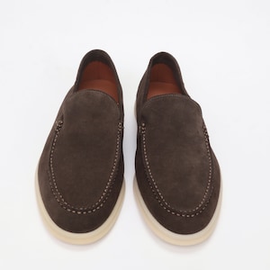 Op de afbeelding: Een paar bruine su&egrave;de loafers met een witte rubberen zool. De loafers hebben een gestikt detail rond de rand van de schoen.