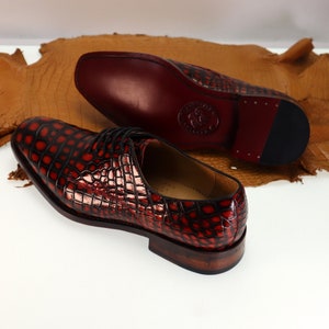 May include: A pair of red leather dress shoes with a crocodile pattern. The shoes have a lace-up closure and a brown leather sole.