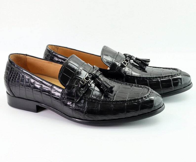 May include: A pair of black leather loafers with a crocodile pattern and tassel accents. The loafers have a pointed toe and a low heel.