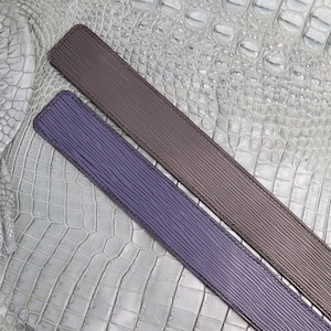 Puede incluir: Dos cinturones de cuero, uno marrón y otro morado, se colocan sobre un fondo de piel de cocodrilo gris.