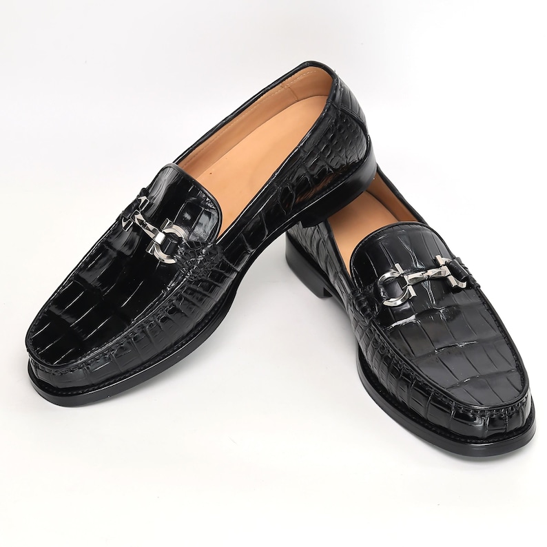 Op de afbeelding: Paar zwarte leren loafers met een krokodillenleertextuur. Elke schoen heeft een zilverkleurige metalen gesp op de wreef. De schoenen hebben een klassiek instapontwerp met een lage hak.