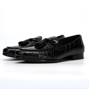 May include: A pair of black leather loafers with a crocodile texture. Each shoe features a tassel detail on the vamp. The shoes have a low heel and a glossy finish, suitable for formal occasions.