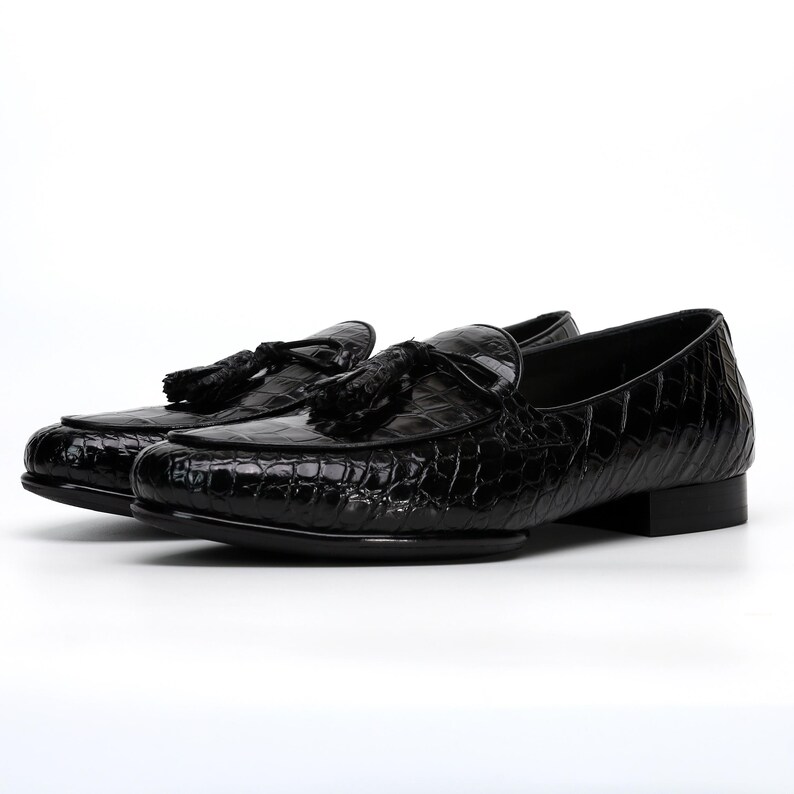 May include: A pair of black leather loafers with a crocodile texture. Each shoe features a tassel detail on the vamp. The shoes have a low heel and a glossy finish, suitable for formal occasions.