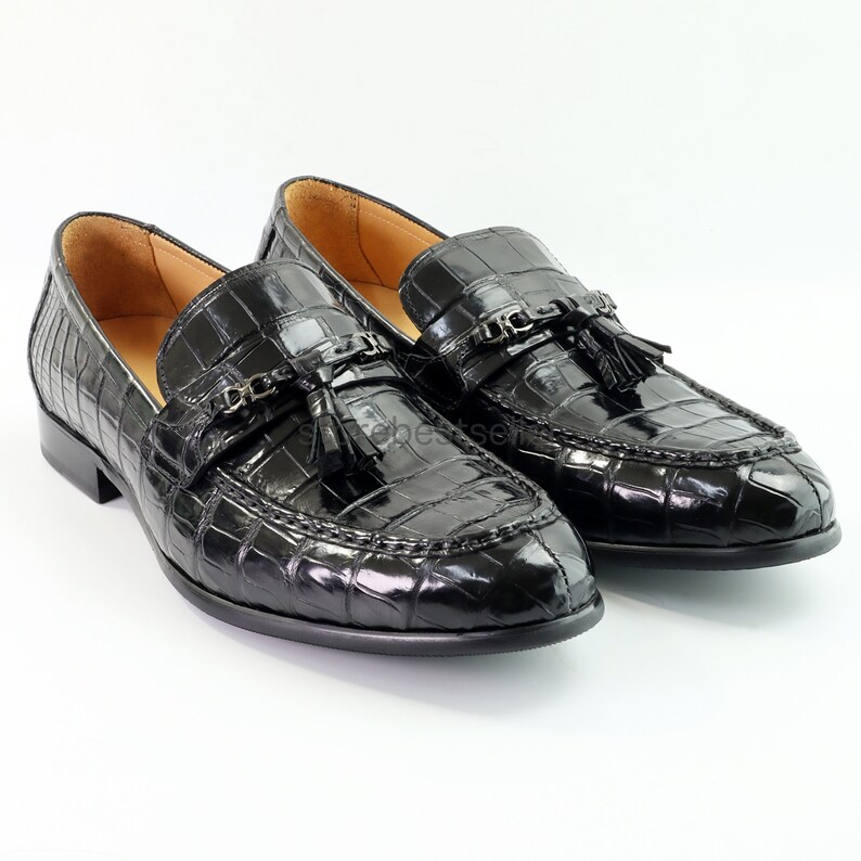 May include: A pair of black leather loafers with a crocodile pattern and tassel accents. The loafers have a pointed toe and a low heel.