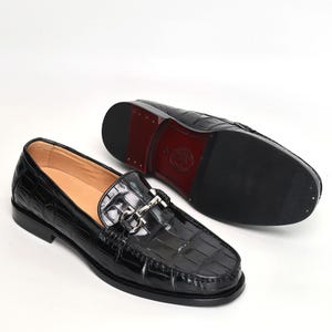 Op de afbeelding: Zwarte leren loafers met een krokodillenpatroon. De schoenen hebben een zilverkleurige metalen gesp en een rode zool. Het ontwerp is klassiek en elegant, geschikt voor formele of semi-formele gelegenheden.