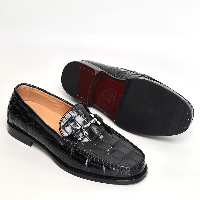 Op de afbeelding: Zwarte leren loafers met een krokodillenpatroon. De schoenen hebben een zilverkleurige metalen gesp en een rode zool. Het ontwerp is klassiek en elegant, geschikt voor formele of semi-formele gelegenheden.