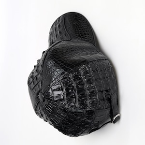 May include: Black leather cap with a textured crocodile pattern. The cap features a rounded crown, a small peak, and a strap with a silver buckle. The cap is made of shiny, high-quality leather.