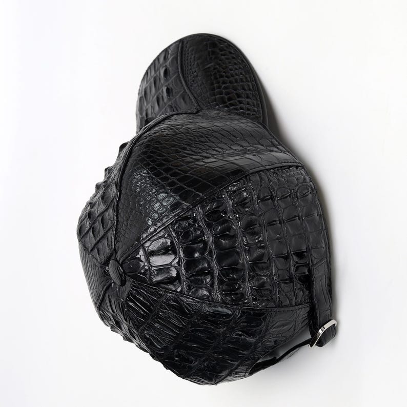 May include: Black leather cap with a textured crocodile pattern. The cap features a rounded crown, a small peak, and a strap with a silver buckle. The cap is made of shiny, high-quality leather.