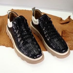 Puede incluir: Un par de zapatillas de cuero negro con detalles blancos y un patrón de piel de cocodrilo en relieve. Las zapatillas tienen cierre de cordones y una suela blanca gruesa.