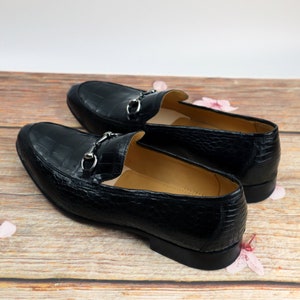 May include: A pair of black leather loafers with a crocodile pattern and silver metal buckles. The loafers are on a wooden surface with pink flowers.