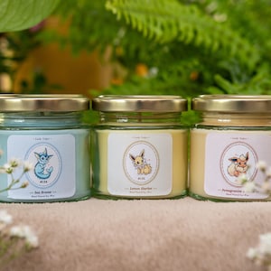 May include: Three glass jar candles with gold lids, each with a different label featuring a cartoon character. The labels read "Sea Breeze", "Lemon Sherbet", and "Pomegranate". The candles are surrounded by greenery and white flowers.