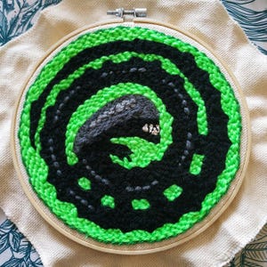 May include: Handmade embroidery art featuring a creature design in a wooden hoop. The artwork showcases a black and green spiral pattern with a gray creature in the center. The embroidery is set on a beige fabric.