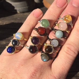 May include: A collection of 12 rings featuring different colored gemstones wrapped in gold wire. The gemstones include amethyst, carnelian, aquamarine, labradorite, tiger's eye, moonstone, lapis lazuli, hematite, rose quartz, citrine, and aventurine.
